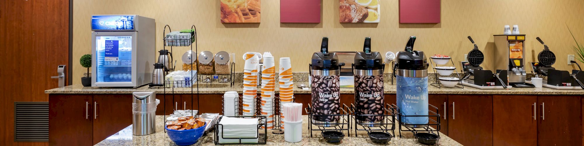 A breakfast buffet featuring coffee dispensers, cereal dispensers, a drink fridge, and various breakfast items on a countertop in a well-lit room.