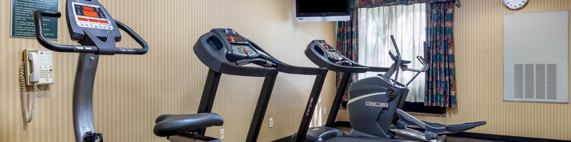 A small gym room with exercise equipment, including a stationary bike, two treadmills, a wall clock, and a wall-mounted TV.