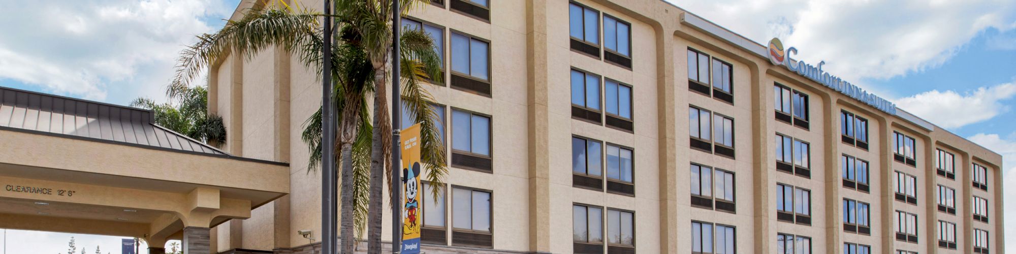 The image shows a multi-story hotel with a parking lot filled with cars and palm trees around the building.