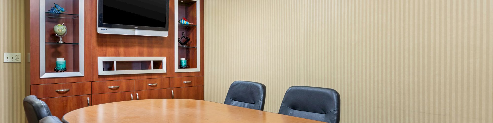 The image shows a conference room with a large oval table and six chairs. A flat-screen TV is mounted on a wooden cabinet.