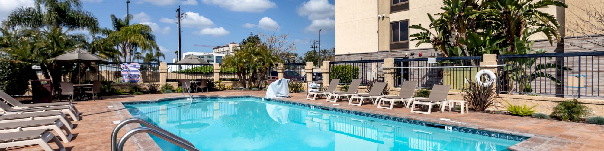 A swimming pool with lounge chairs and umbrellas is surrounded by palm trees and a tall building under a blue sky with clouds.