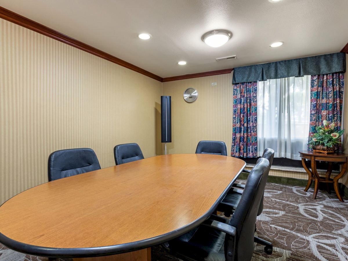 A conference room with a wooden table, six chairs, a window with floral curtains, and a small side table with a floral arrangement.