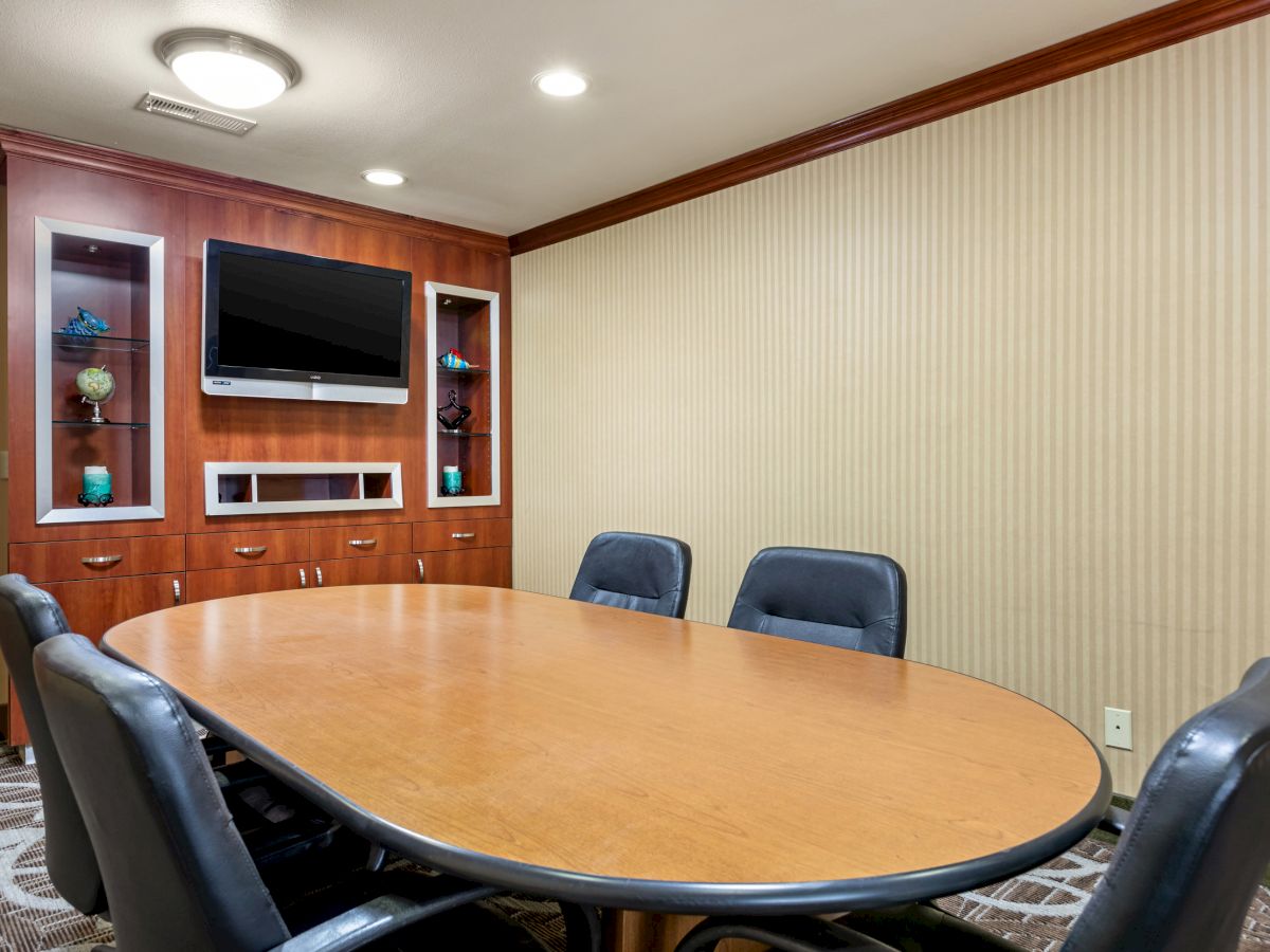 The image shows a conference room with a large oval table, six black chairs, a television mounted on a wooden panel, and various decorative items.