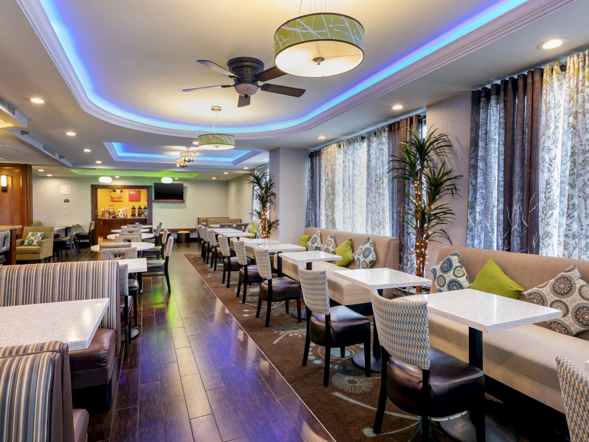 This image shows a modern dining area with stylish chairs, tables, decorative cushions, ceiling lights, and a TV, featuring greenery and blue LED lighting.