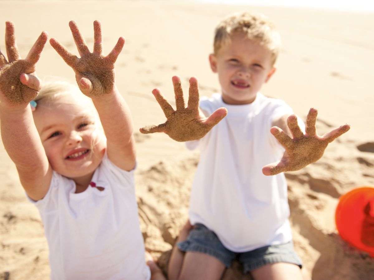 Two children playing on the beach, smiling and showing their sandy hands. There is a red bucket on the ground beside them.