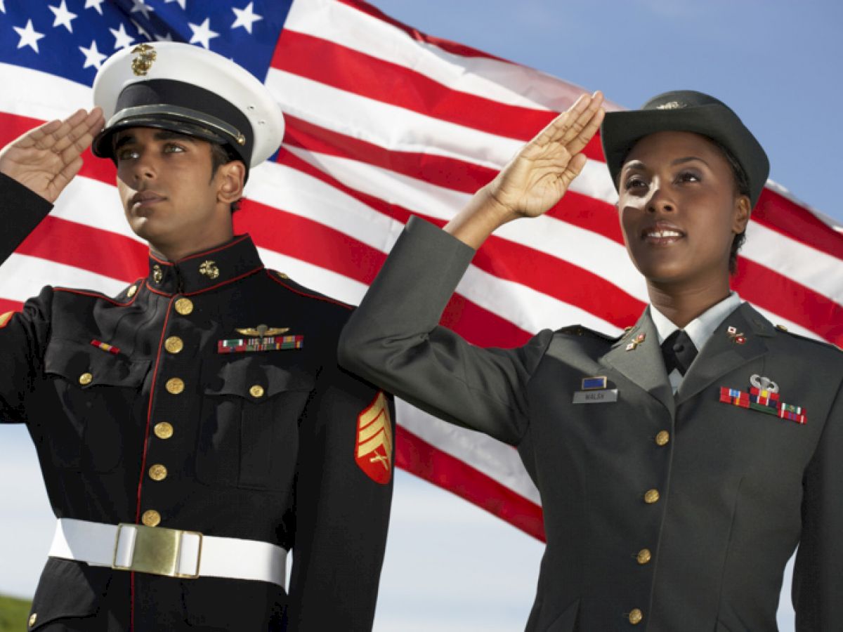 Two military personnel salute in front of an American flag, one in a Marine Corps uniform and the other in an Army uniform, under a clear sky.