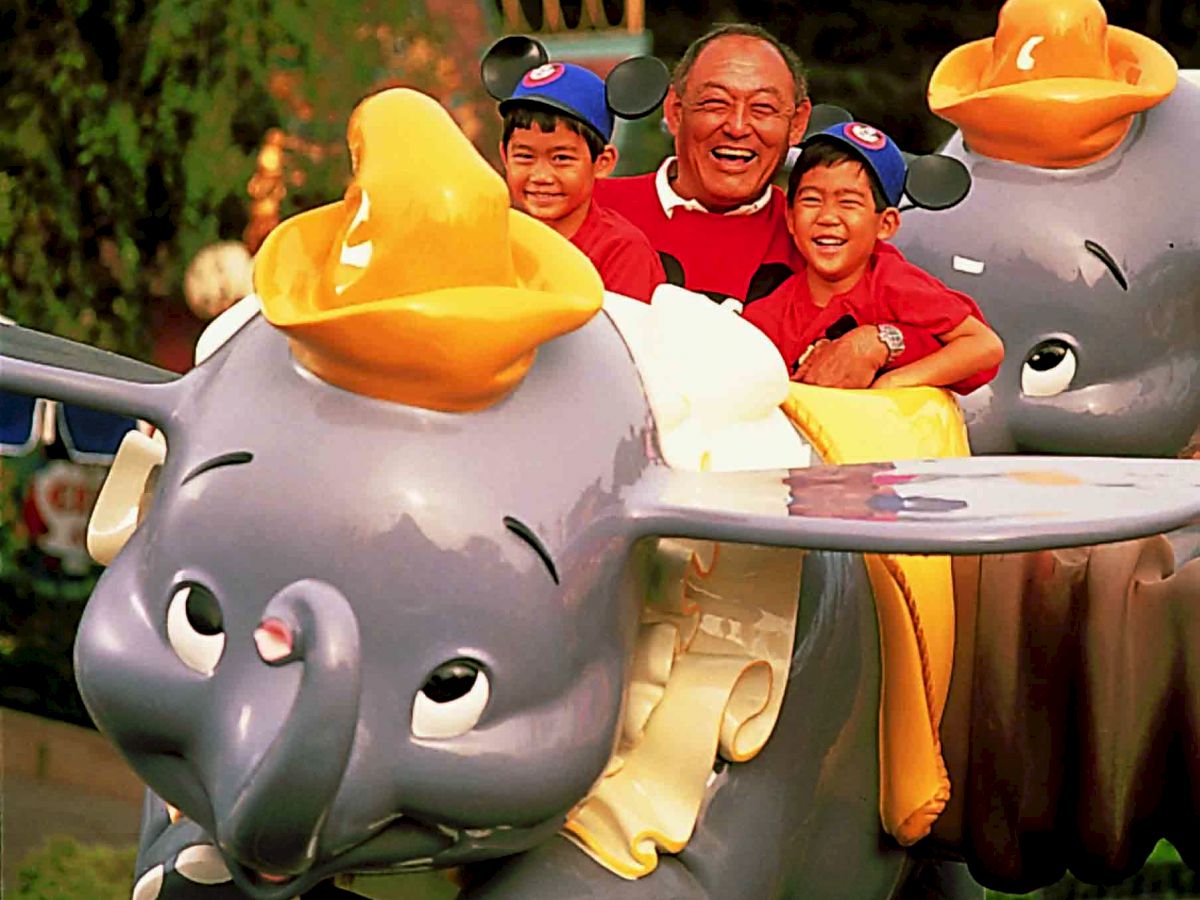 Three people enjoying a ride on Dumbo the Flying Elephant at a theme park, with two children wearing Mickey Mouse ears hats.