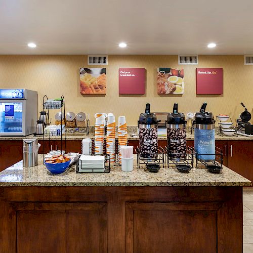 A breakfast buffet setup with coffee dispensers, cereal dispensers, cups, an ice machine, and a variety of food options on the counter.