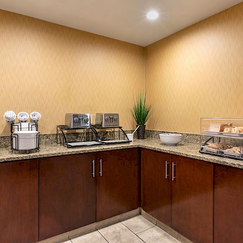 The image shows a hotel breakfast buffet area with coffee dispensers, a plant, covered trays with pastries, condiments, granola, and cabinets.