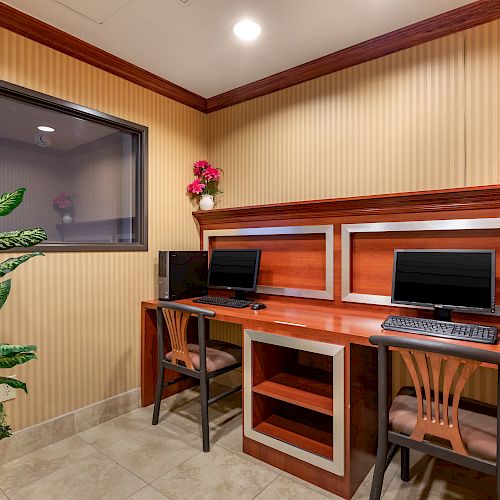 A small office space with two desks, each with a computer, chairs, a plant, and wall decor with flowers, under indoor lighting.