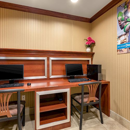 A small workspace with two computer stations, wooden desks, chairs, flowers, and a colorful poster on the wall featuring a theme park advertisement.