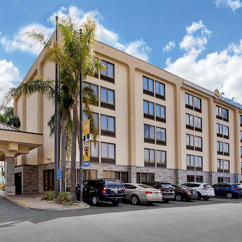 This image shows a multi-story hotel building with a parking lot and several parked cars in front of it.