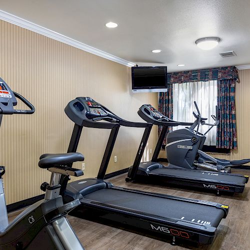 The image shows a small gym room with exercise equipment, including two treadmills, a stationary bike, and an elliptical machine, under ceiling lights.