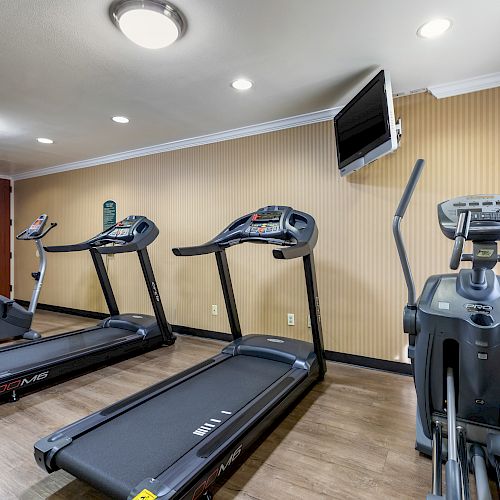 The image shows a fitness room with three treadmills, an elliptical machine, a TV mounted on the wall, and a stack of clean towels.