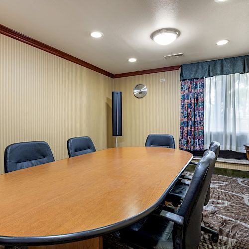 A small conference room with a large oval table, six chairs, floral curtains, a side table with a plant, and wall clock.