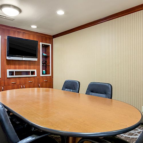 A conference room with a long oval table, six chairs, a wall-mounted TV, and built-in wooden shelves with decorative items.