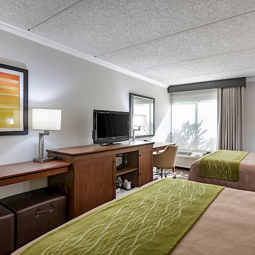 The image shows a hotel room with two beds, a TV on a wooden stand, a desk with a chair, two lamps, and a window with curtains.