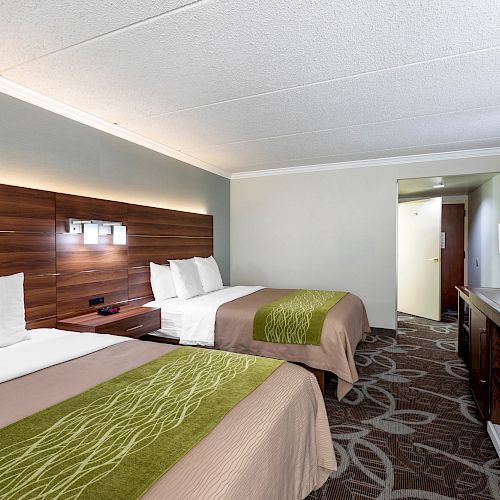 A hotel room features two double beds with green runners, a flat-screen TV, a desk, and a lamp. The design is modern and clean, with neutral tones.