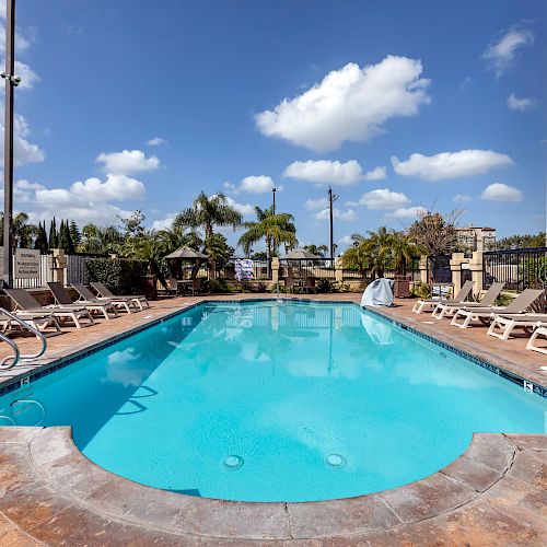 An outdoor swimming pool with lounge chairs on both sides, surrounded by a fence. Palm trees and a building can be seen in the background.