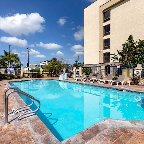 An outdoor pool area with lounge chairs, surrounded by a fence, with palm trees and a multi-story building in the background under a blue sky.