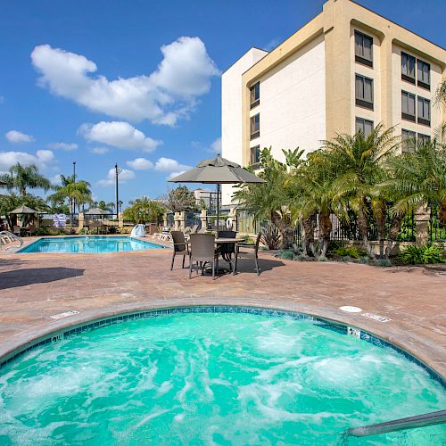 A hotel pool area with a hot tub in the foreground, chairs and tables with umbrellas, and a larger pool and building in the background.