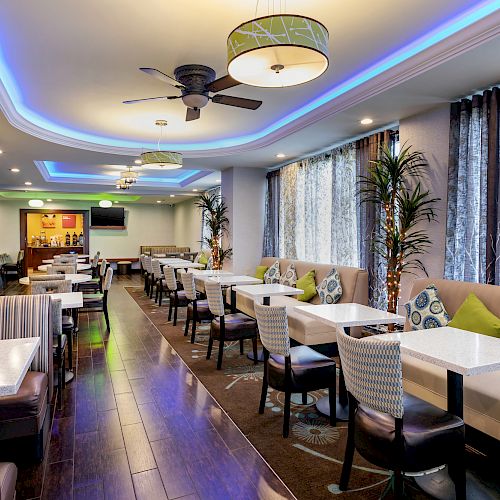 A brightly lit modern restaurant interior with striped booth seating, white tables, cushioned chairs, ceiling fans, and potted plants next to large windows.