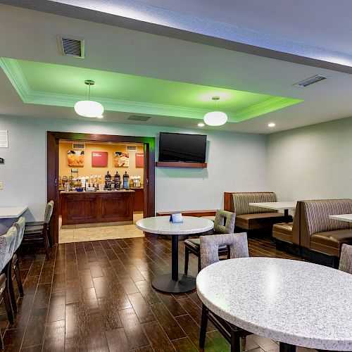 A dining area with tables, chairs, a TV, and booths, featuring a brightly lit green ceiling and a small buffet zone near the back.