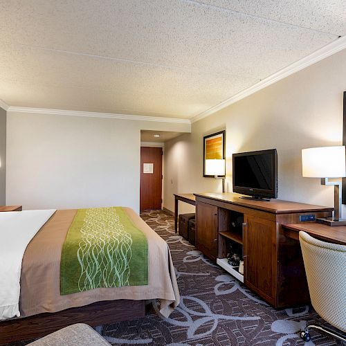 This image shows a hotel room with a neatly made bed, a desk with a chair, a flat-screen TV on a stand, and modern decor.