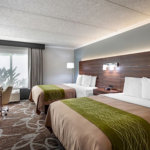 The image shows a hotel room with two double beds, a wooden headboard, a TV on a dresser, a desk with a chair, and a window with curtains.