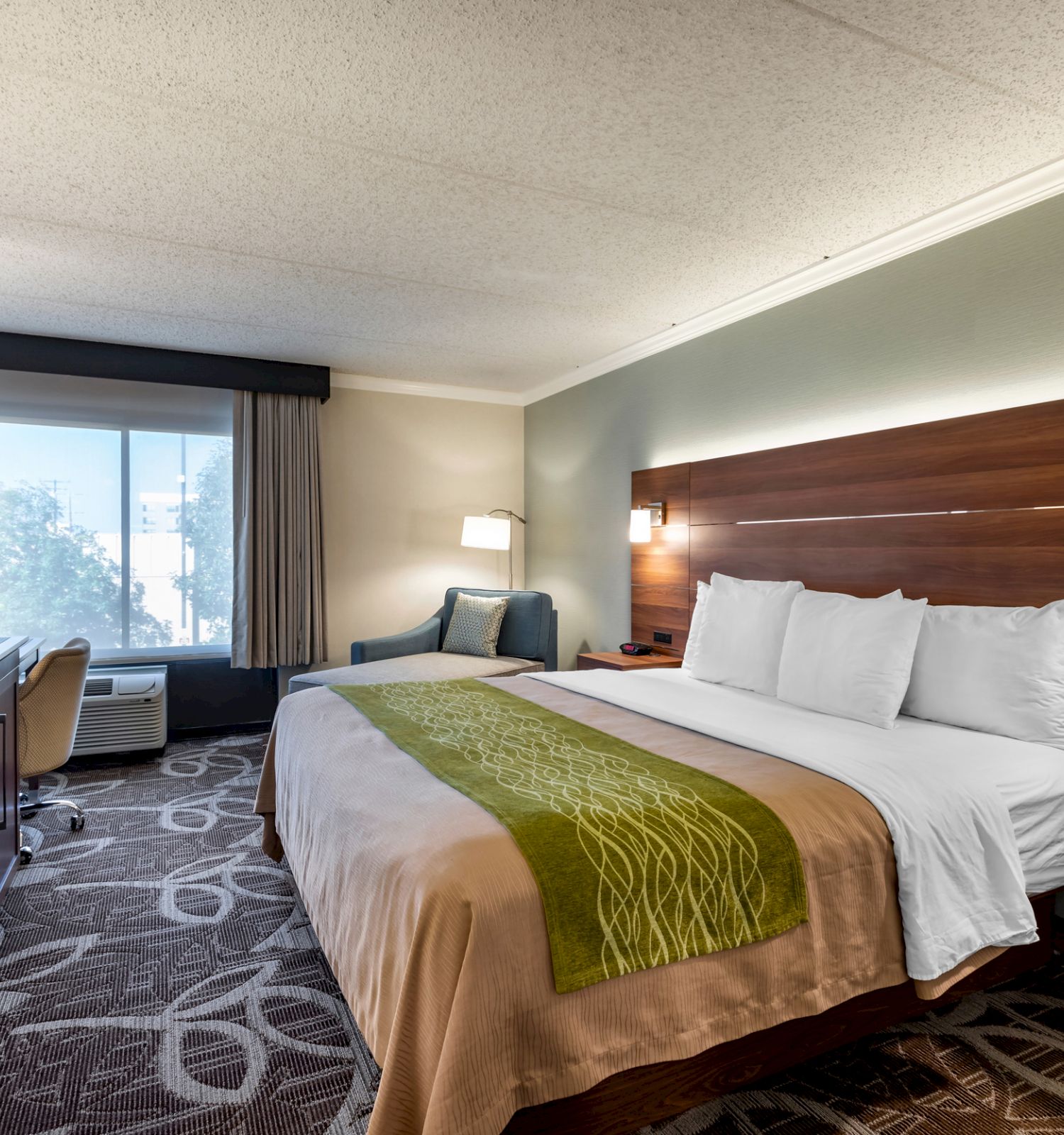 A hotel room with a king-size bed, a TV on a wooden desk, a chair, a window with curtains, and wall lamps above the bed.