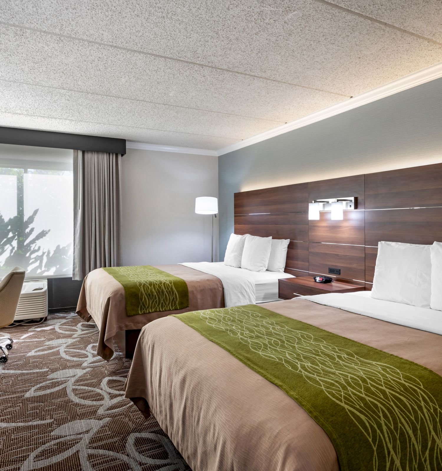 A hotel room with two beds, a TV, a desk with a chair, and a window with curtains. Brown and green bedding with patterned carpet.