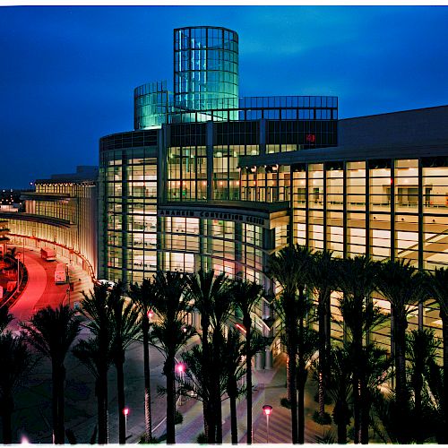 The image shows a modern glass and steel building at dusk, with an illuminated sky and palm trees in the foreground.