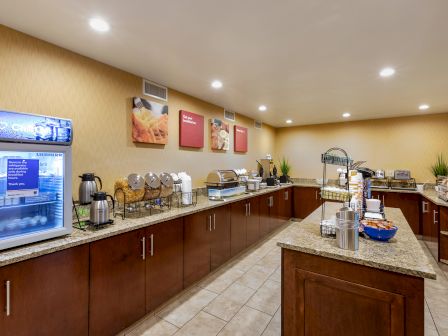 This image shows a well-lit breakfast buffet area with a variety of food and beverages, including a display fridge, coffee makers, and buffet counters.