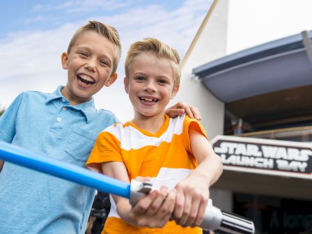 Two kids, one in a blue shirt and the other in an orange one, are holding a blue lightsaber in front of a 