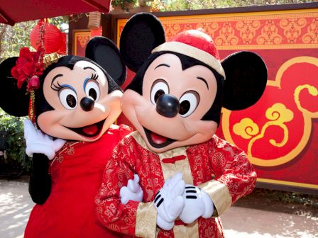 Two costumed characters, resembling mice, are dressed in traditional red and gold attire, posing in front of a festive backdrop.