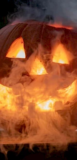 A carved pumpkin jack-o'-lantern, illuminated from within and surrounded by smoke, is shown in the image.