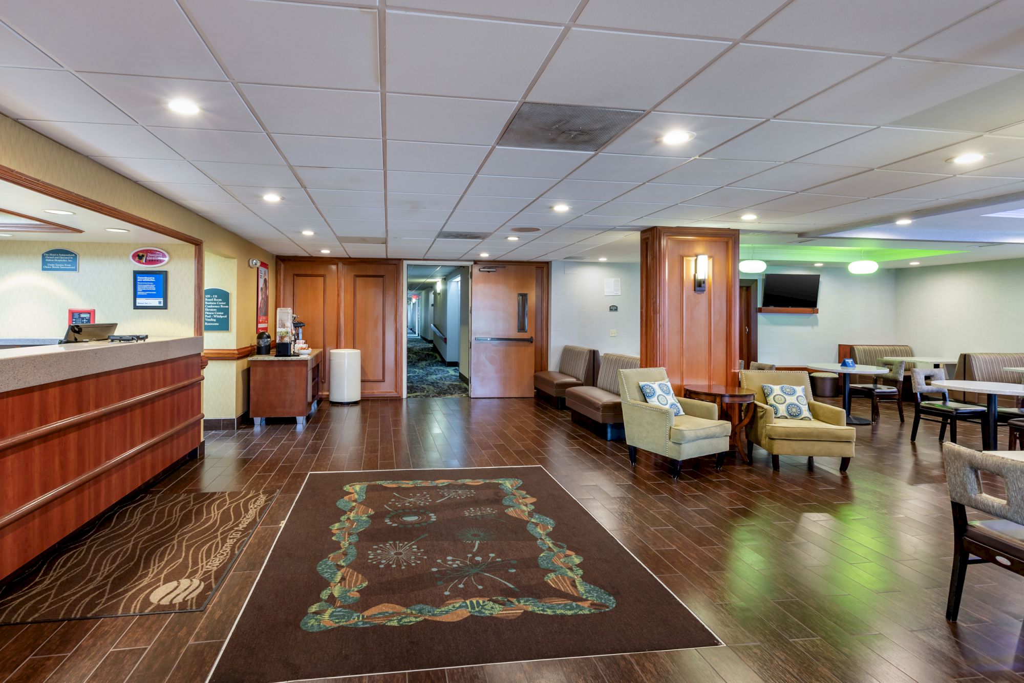 This image shows a hotel lobby with a front desk, seating areas, a hallway entrance, and a television on the wall.