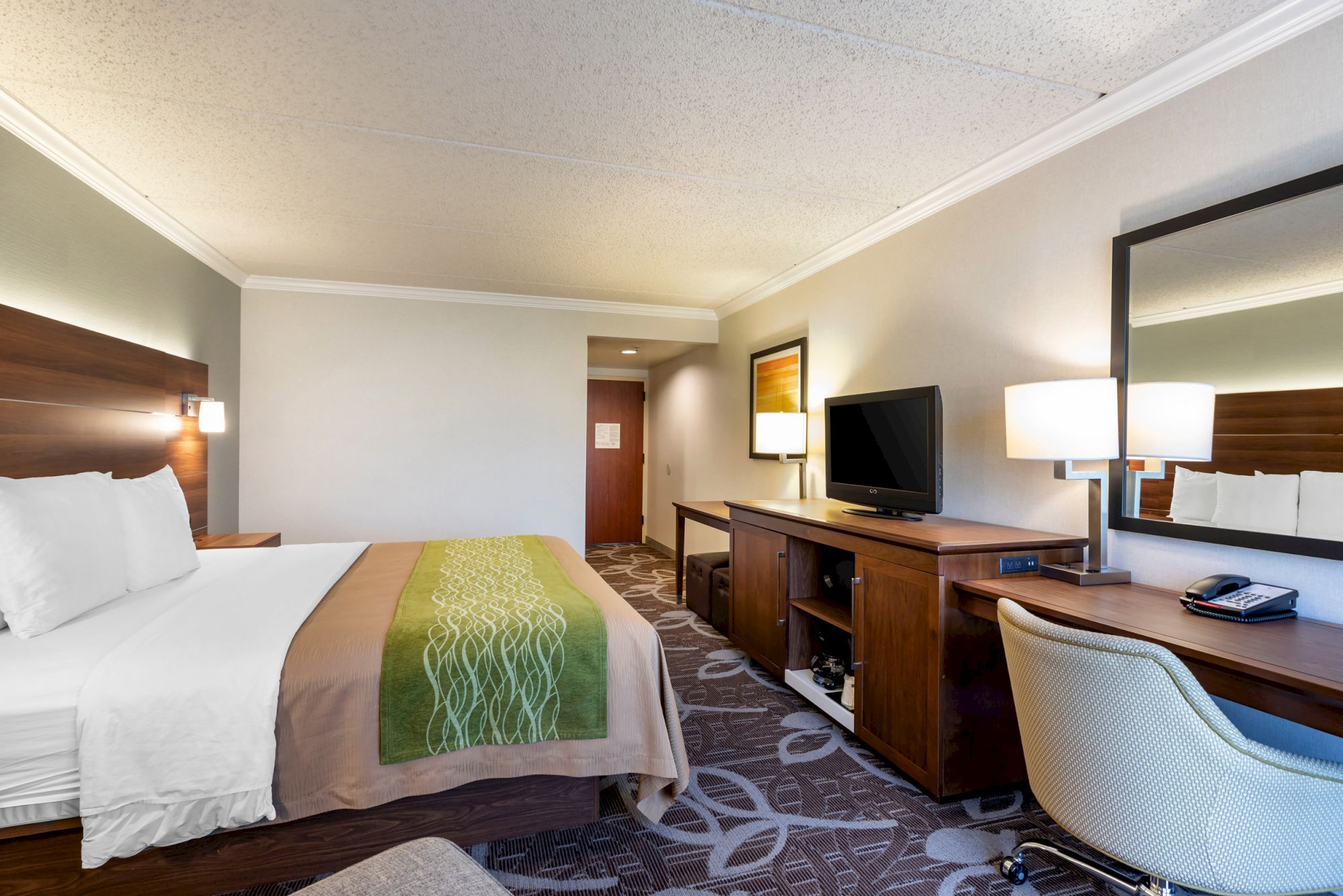 A hotel room with a king-sized bed, TV, desk and chair, mirror, lamps, and modern decor. The room looks clean and comfortable, featuring neutral colors.