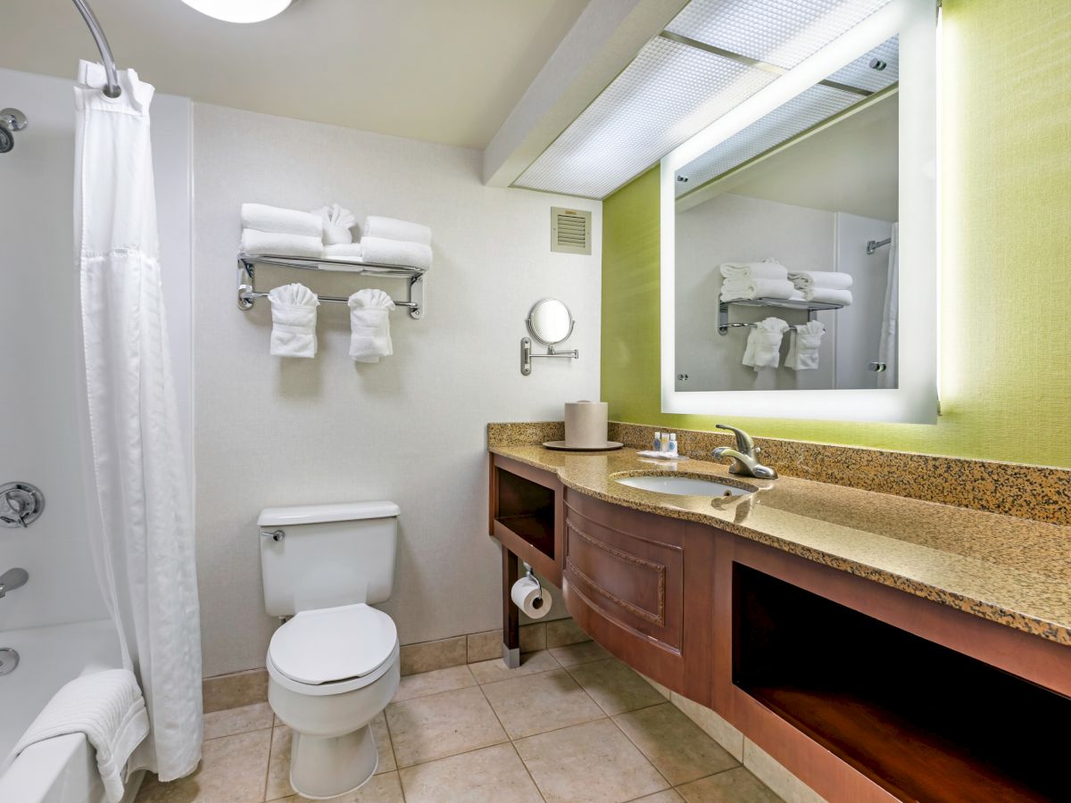 This image shows a bathroom with a bathtub, toilet, vanity with a sink, towels, mirror with lights, and tiled floors.